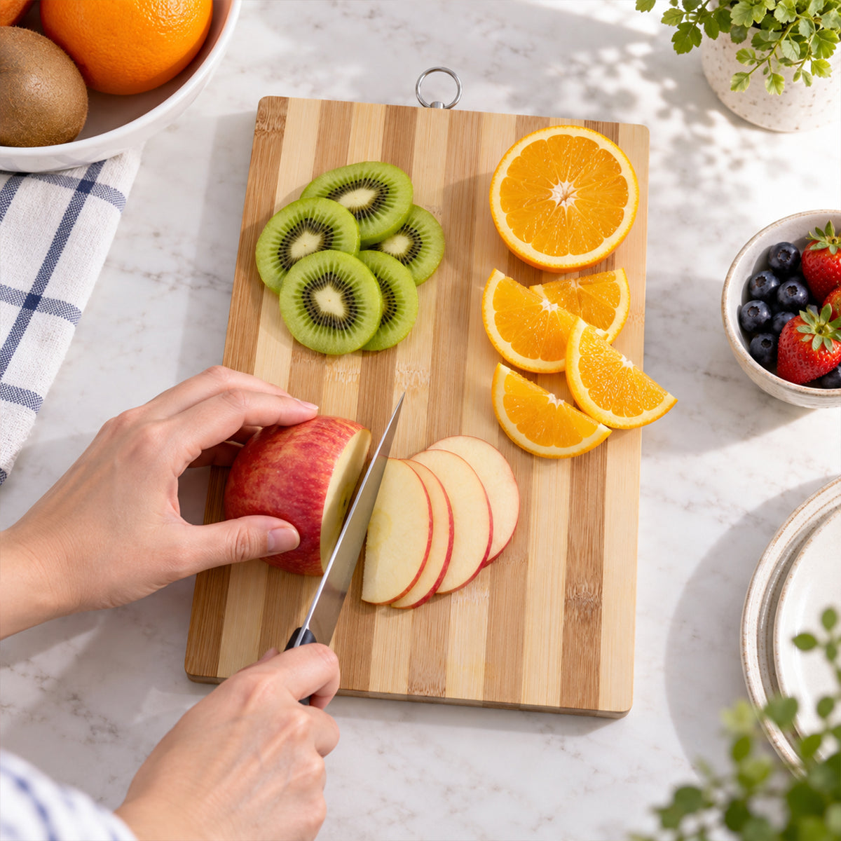 A person slices an apple on a striped bamboo board, surrounded by kiwi, orange, and berries on a white counter.