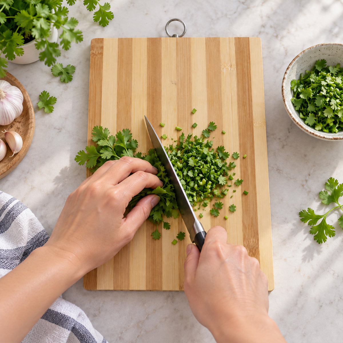 A person's hands chop fresh cilantro on a striped bamboo cutting board, with garlic and a bowl of herbs nearby.