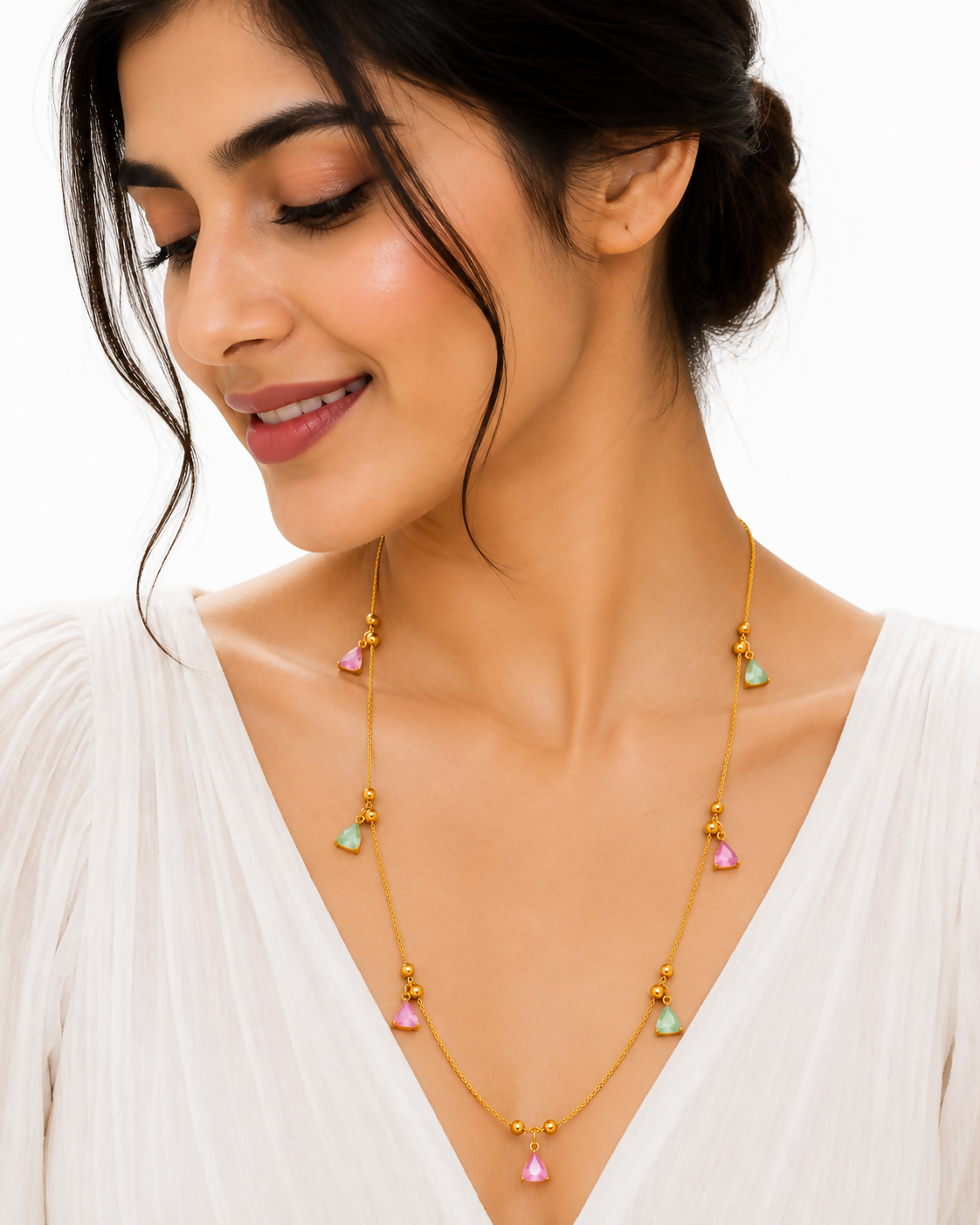 A smiling woman wears a gold-plated necklace with pink and mint green triangular charms over a white dress.