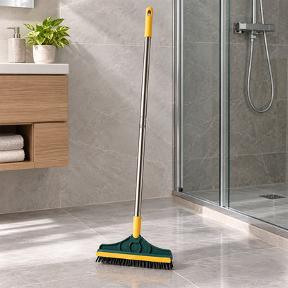 A green and yellow floor cleaning brush with a metal handle stands on a grey tiled bathroom floor, next to a shower and