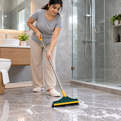 A woman in a grey shirt and beige pants scrubs a tiled bathroom floor with a green and yellow brush, creating white foam.