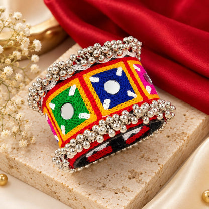 A vibrant embroidered kada bracelet with silver-tone beads and small mirrors rests on a light brown surface.