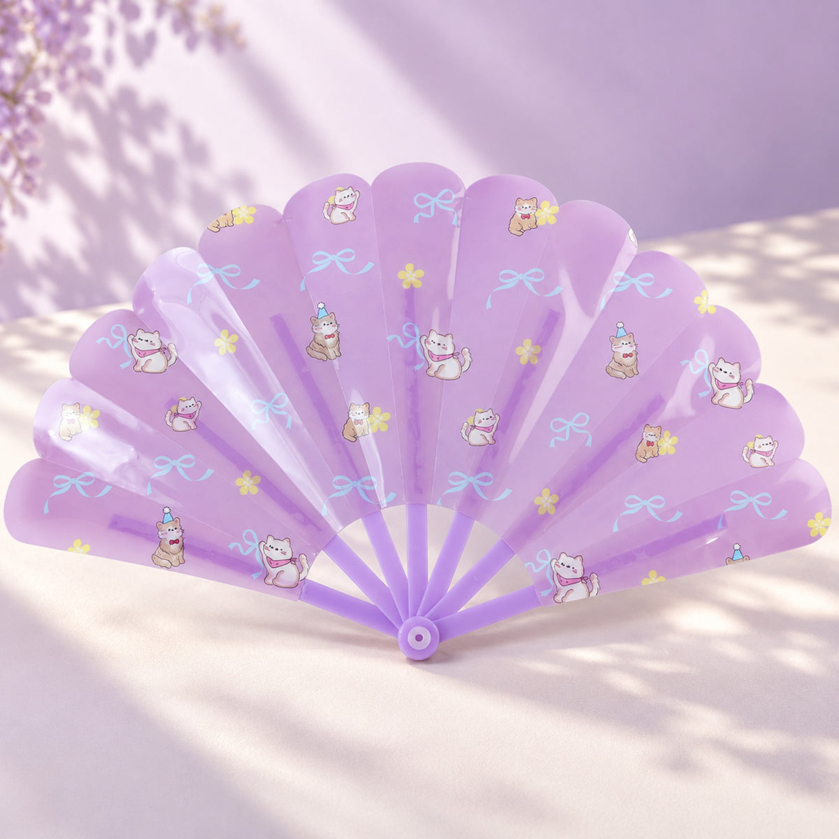 An open purple hand fan with playful cat and bow patterns rests on a light purple surface with shadows.