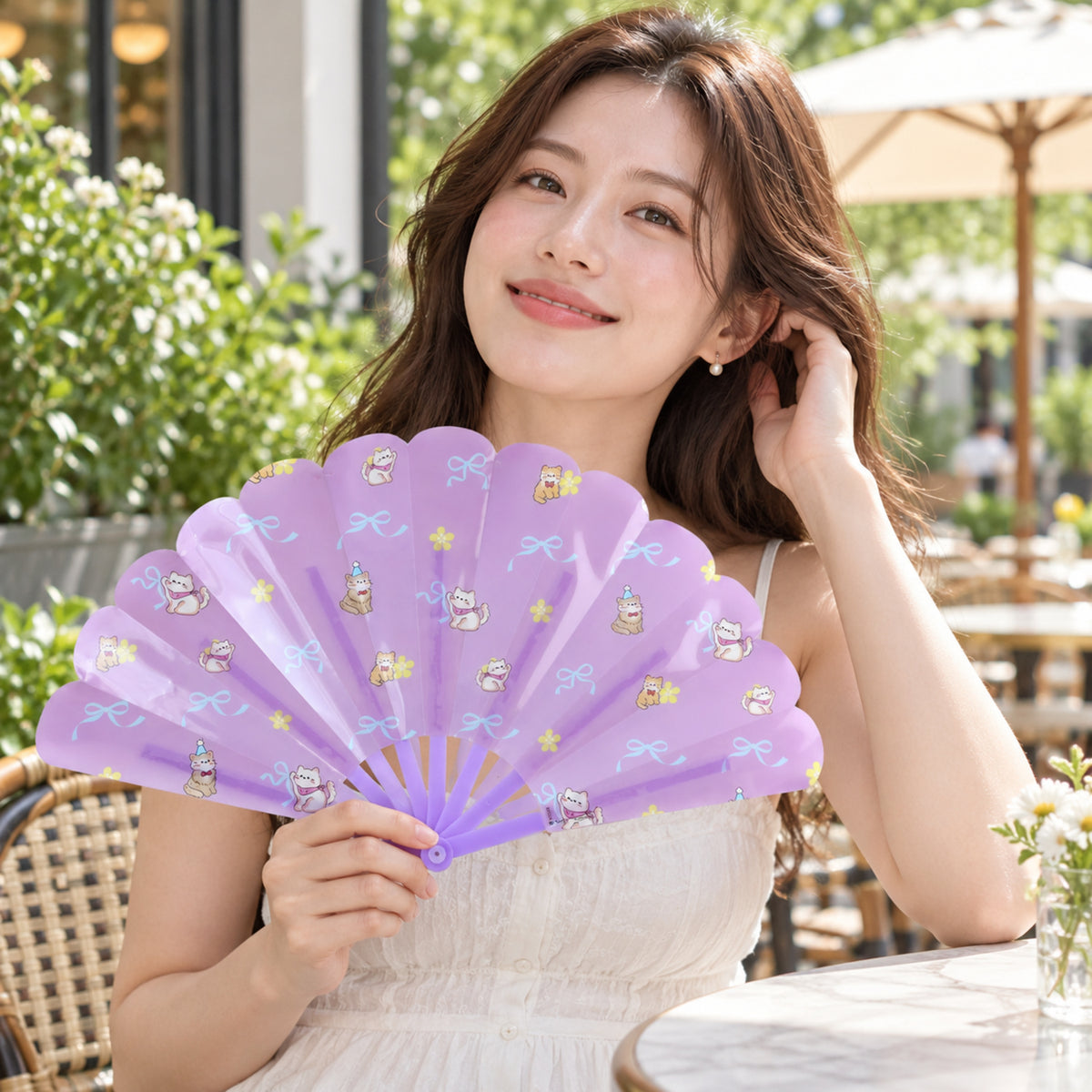 A woman in a white dress holds an open purple fan with cat designs while seated at an outdoor cafe table.
