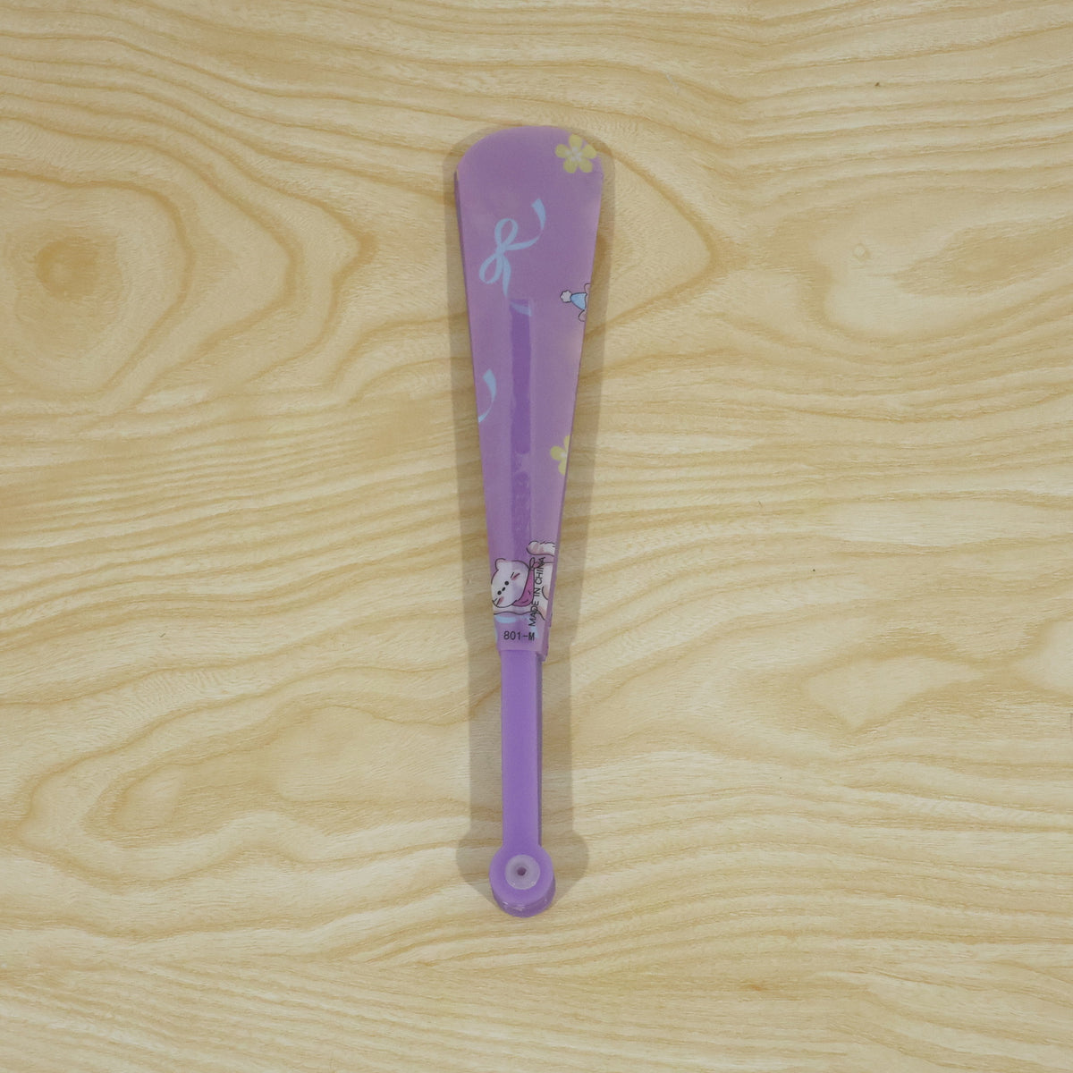 A closed purple folding hand fan with a subtle cat and bow pattern lies vertically on a light wooden surface.