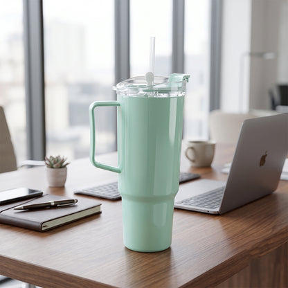 A mint green tumbler with a straw and handle sits on a wooden desk next to a laptop and notebook.
