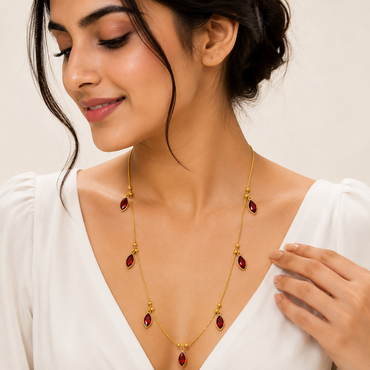 A woman with dark hair wears the gold-plated chain necklace with red crystal pendants over a white top, looking down with a