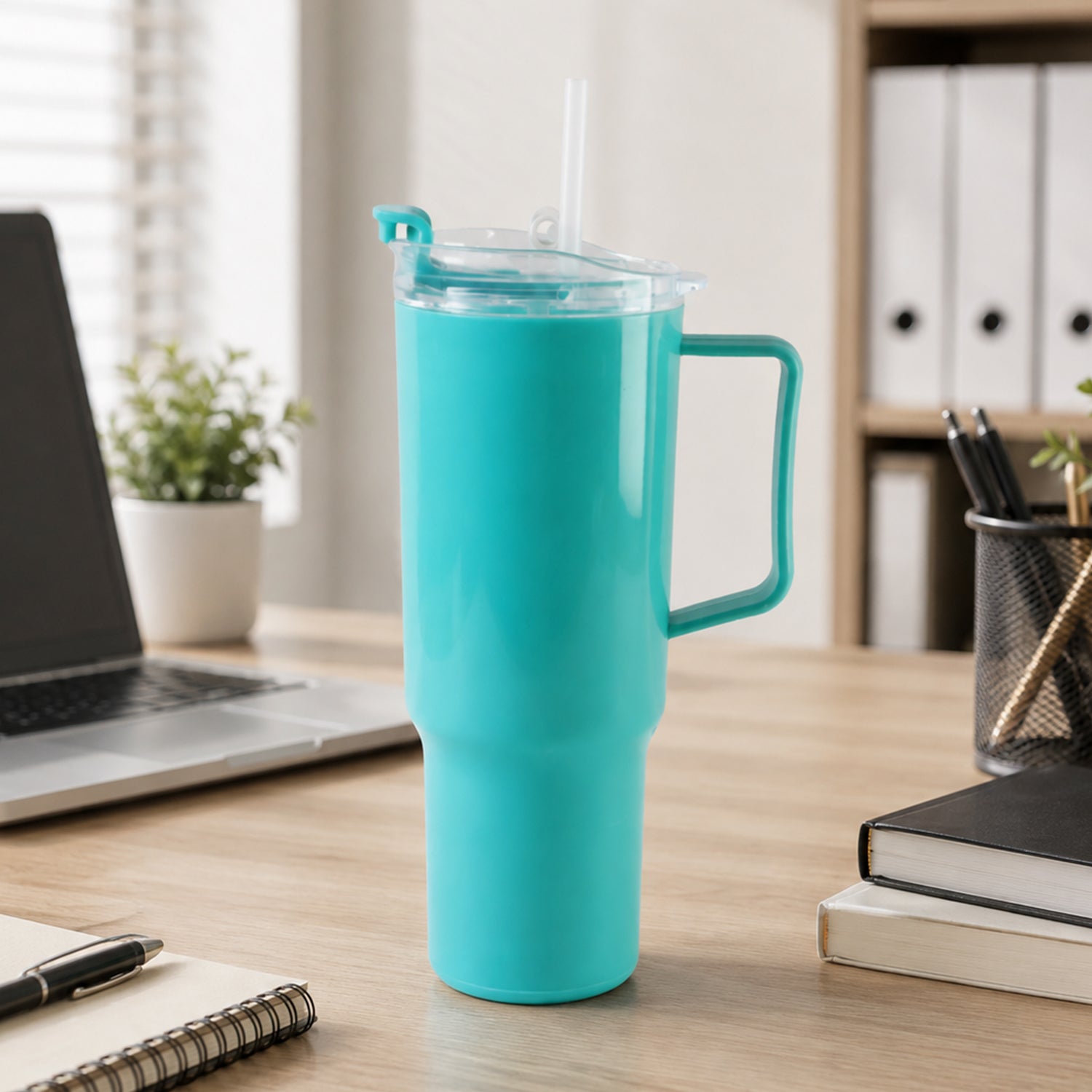 An aqua blue tumbler with a handle, clear lid, and straw sits on a wooden desk next to a laptop and office supplies.