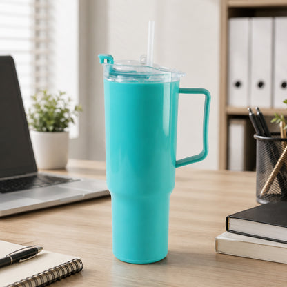 An aqua blue tumbler with a handle, clear lid, and straw sits on a wooden desk next to a laptop and office supplies.