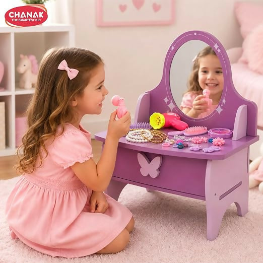 A young girl in a pink dress plays with a toy hair dryer at a purple and pink wooden vanity with a mirror.