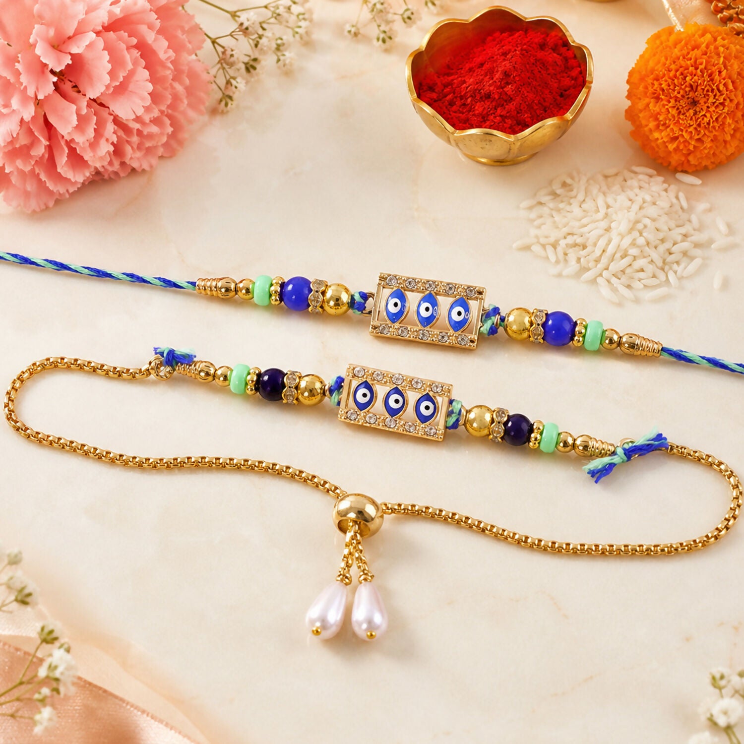 Two blue and gold evil eye rakhis, one a bracelet, on a white surface with pink flowers and rice.