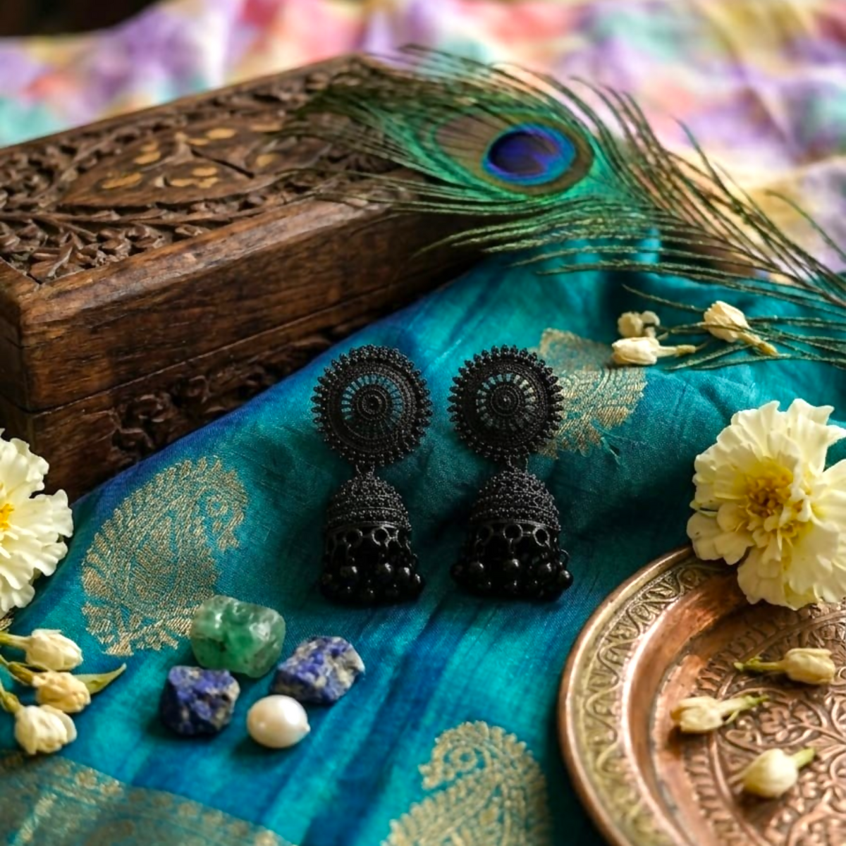 The black oxidised jhumka earrings are displayed on a vibrant blue and gold patterned fabric with decorative elements.
