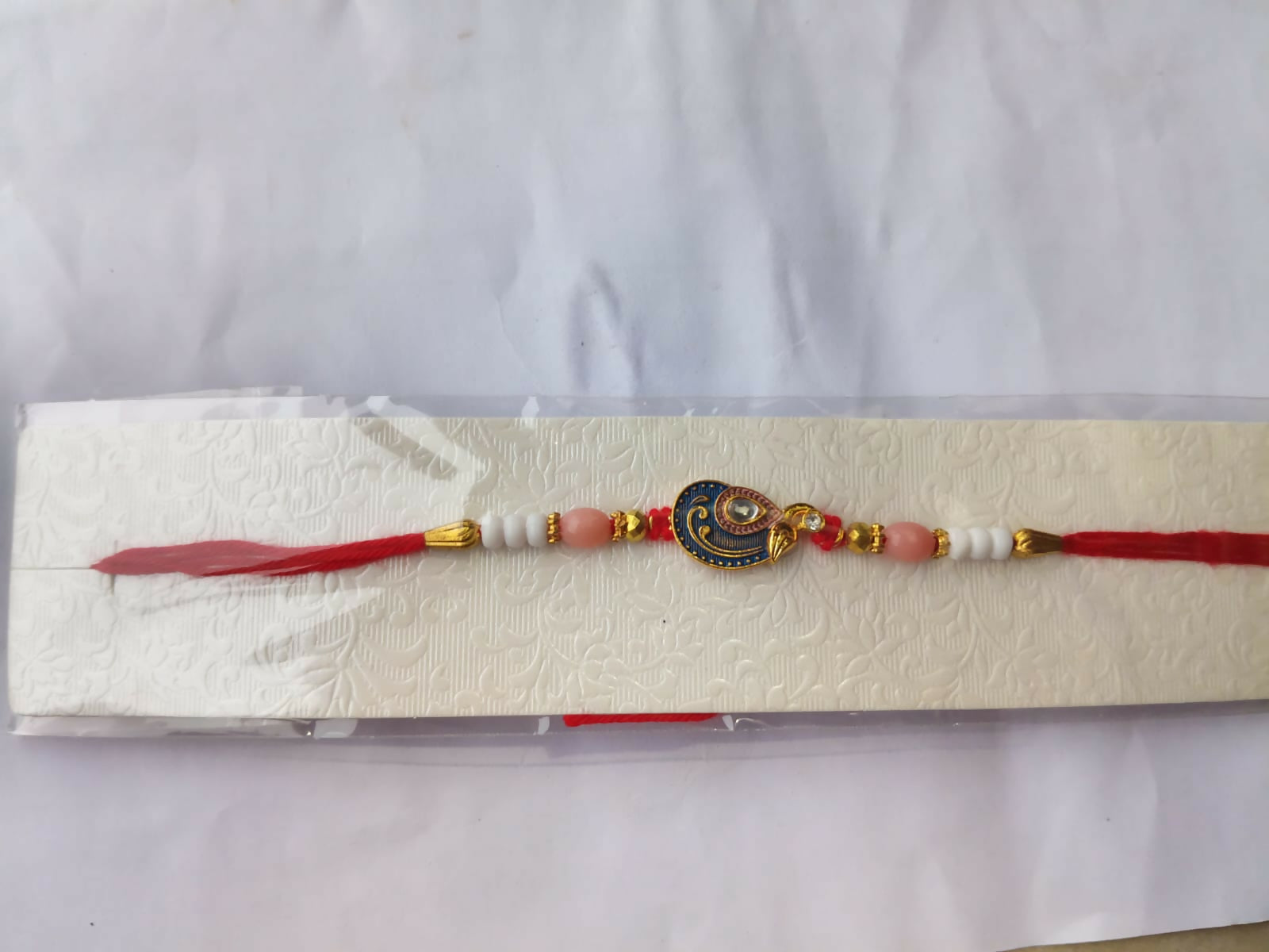 A red, blue, and gold peacock charm bracelet is packaged in clear plastic on a white textured card, against a white