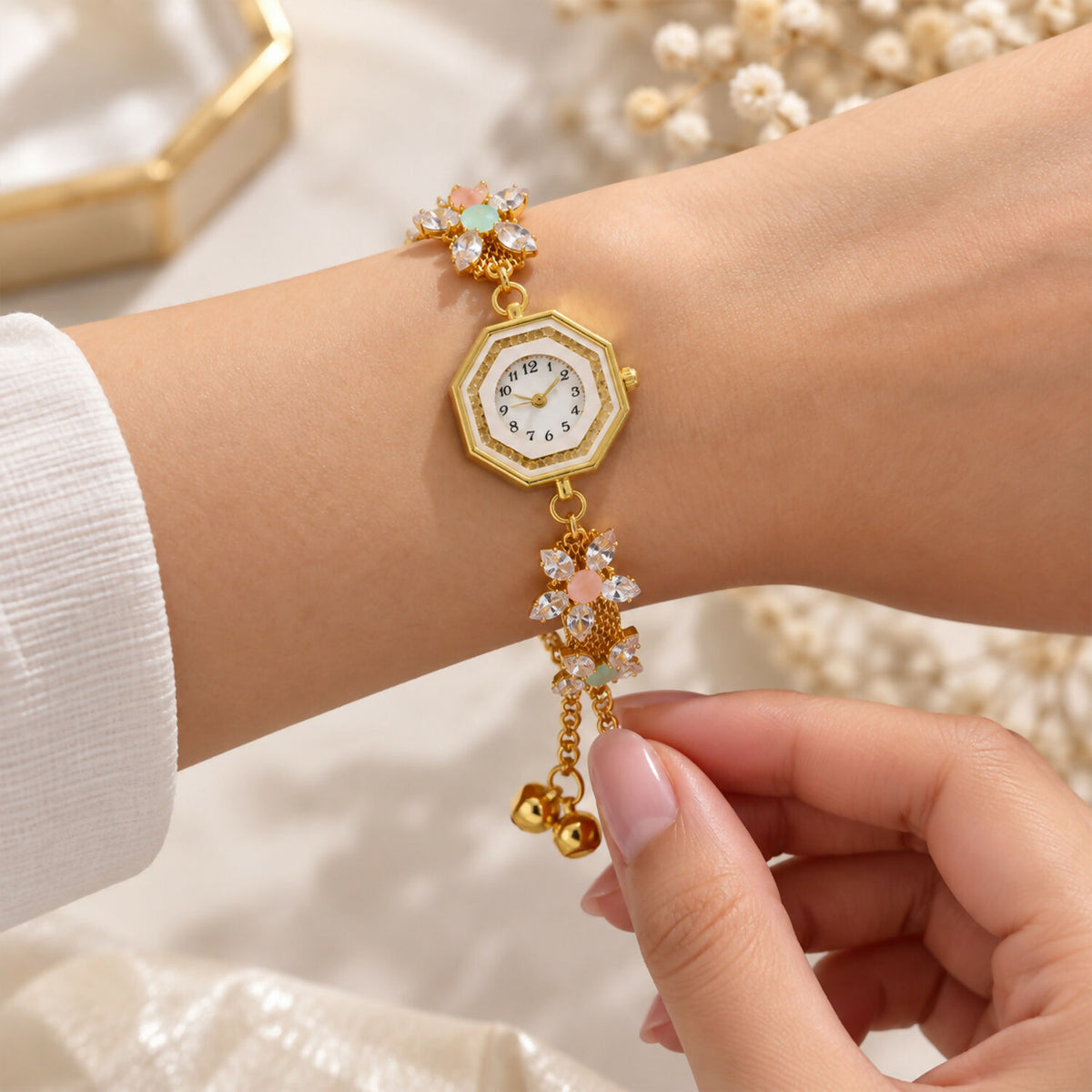 A hand adjusts the gold-tone bracelet watch on a wrist, featuring crystal flowers and decorative bell charms.
