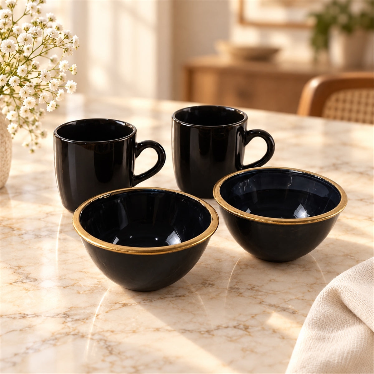 Two black ceramic mugs and two bowls with gold rims sit on a light marble table next to a white flower vase.