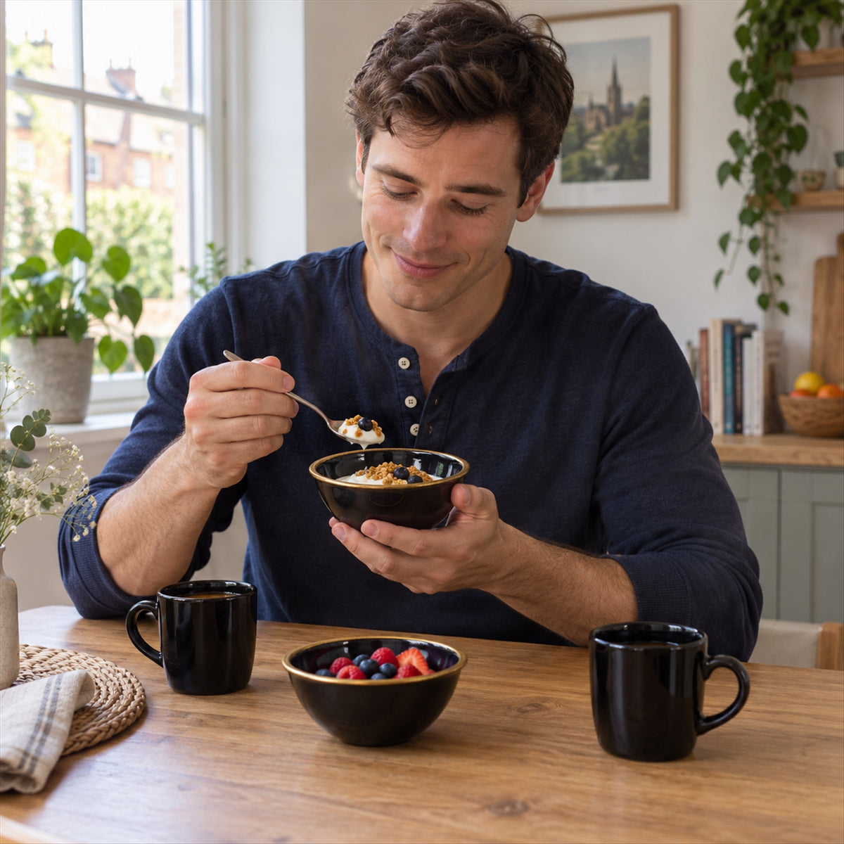 A man in a blue shirt eats cereal from a black ceramic bowl with a gold rim at a wooden table with two mugs.