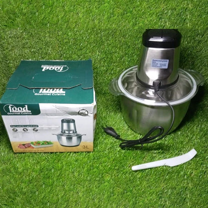 A stainless steel electric food chopper, its power cord, and a green product box resting on artificial grass.