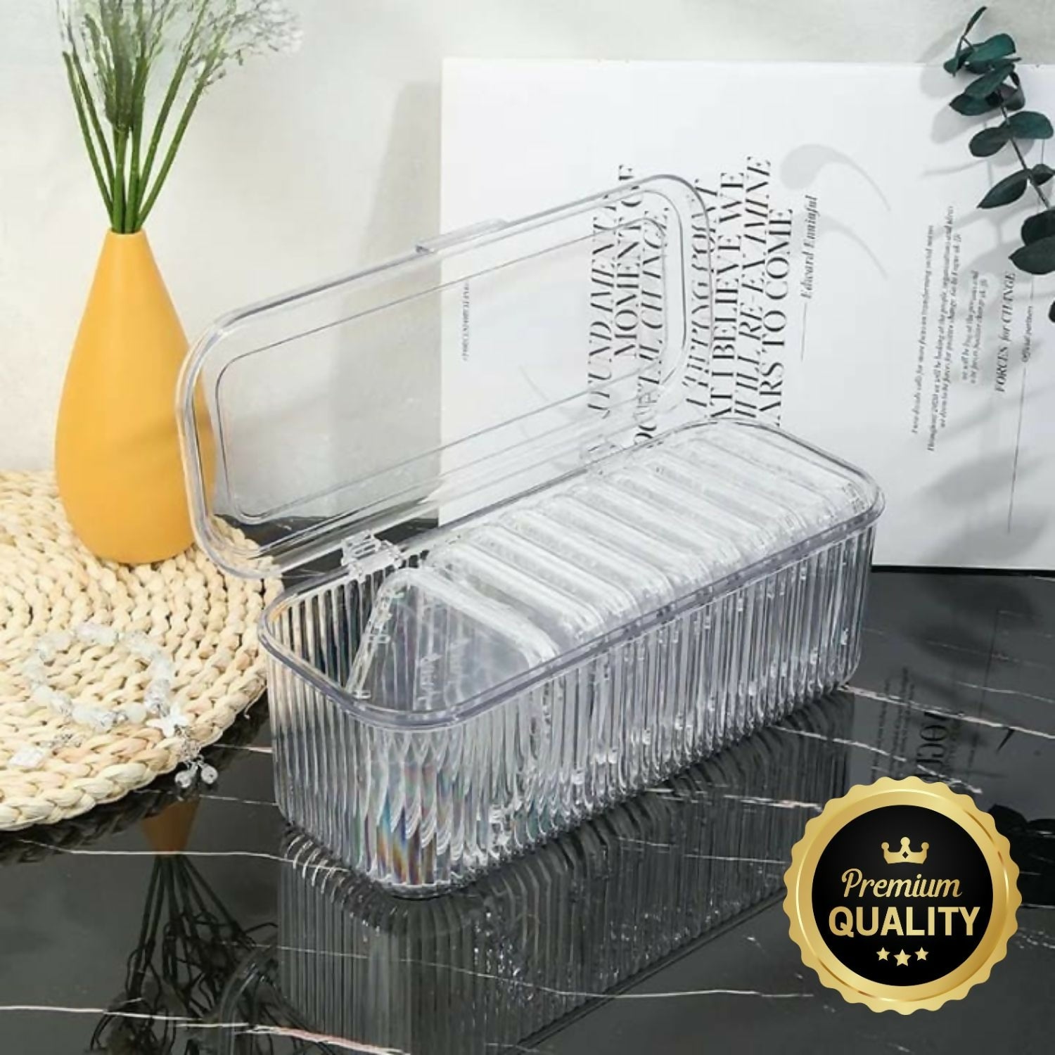 A clear, ribbed plastic storage box with an open flip lid sits on a dark surface next to a yellow vase and white book.