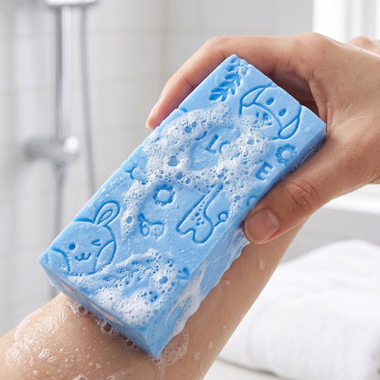 A hand holds a blue exfoliating sponge with animal patterns, scrubbing a leg with soap suds, viewed from a different angle.