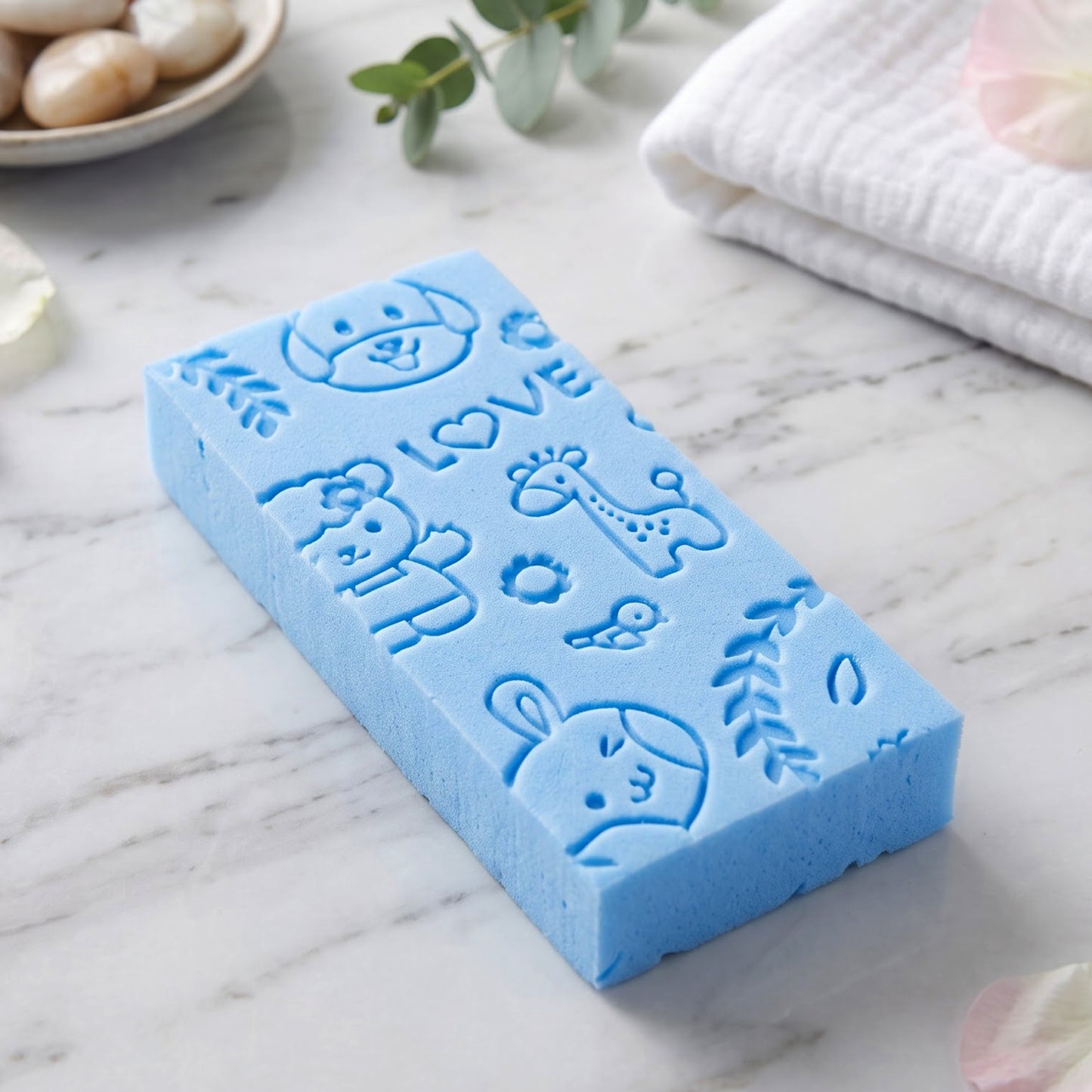 A blue exfoliating sponge with embossed animal patterns rests on a white marble counter with decorative stones and a towel.