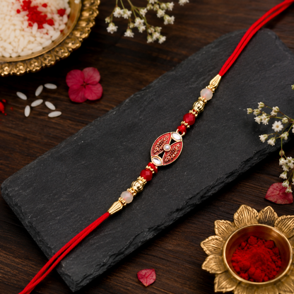 The red and gold beaded bracelet rests on a dark slate surface, with a small gold bowl of red powder in the corner.