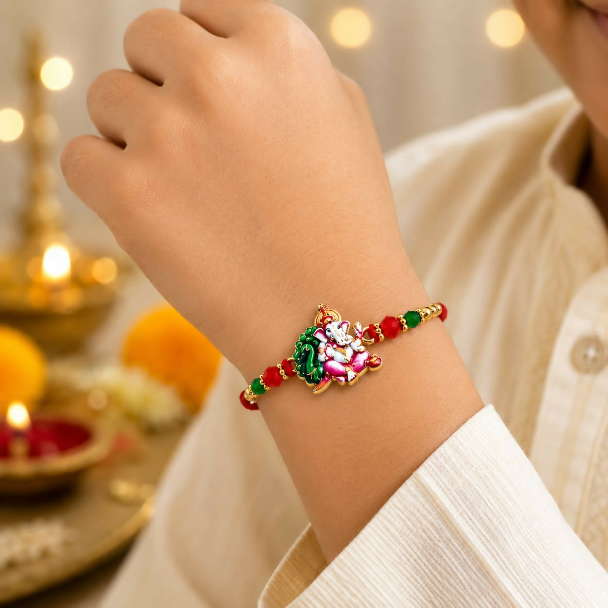 A person's arm wearing a red thread with an enamel Ganesha charm, green and red crystal beads, and golden beads, with