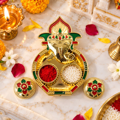 A golden Ganesha-shaped kumkum holder with red powder and white rice on a marble surface, surrounded by flowers and a lit