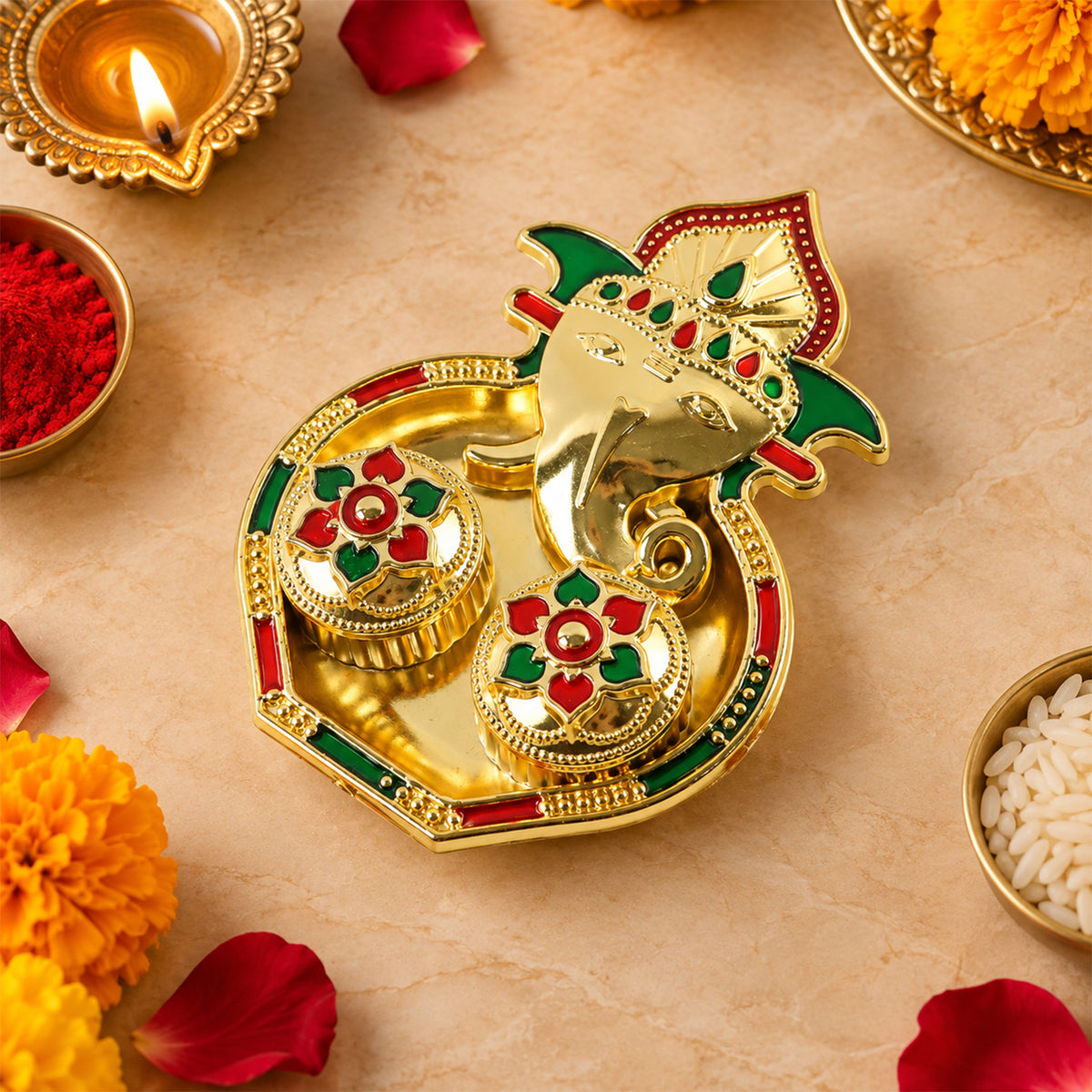 A golden Ganesha-shaped kumkum holder with red and green enamel details, surrounded by marigolds and rose petals.