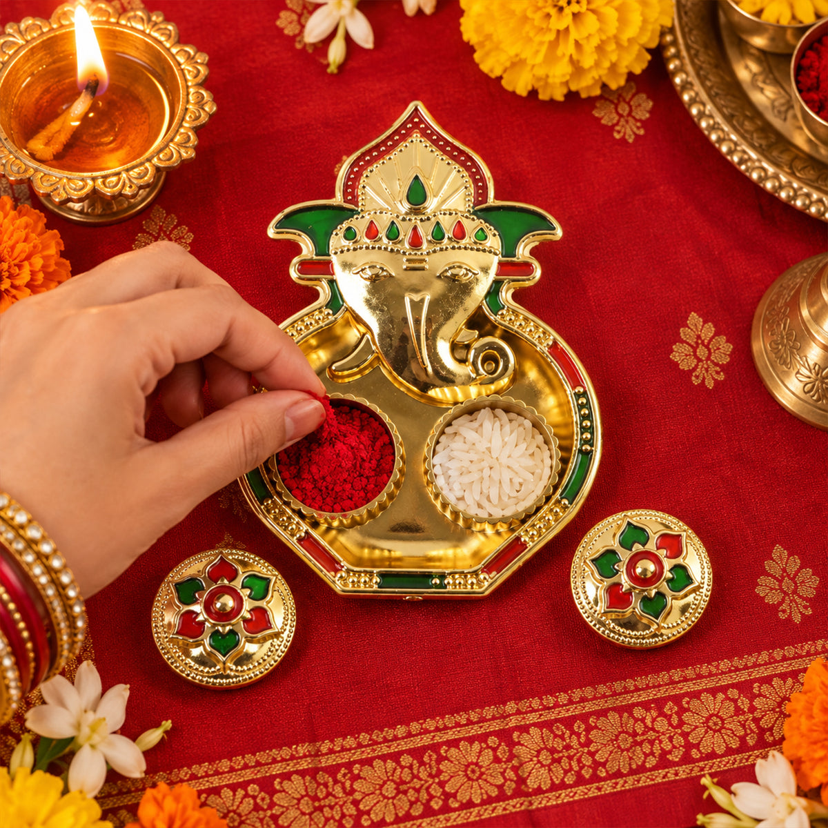 A hand places red kumkum powder into a golden Ganesha-shaped holder on a red patterned fabric.
