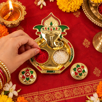 A hand places red kumkum powder into a golden Ganesha-shaped holder on a red patterned fabric.