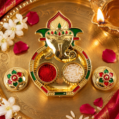 A golden Ganesha-shaped kumkum holder with red powder and white rice, surrounded by white flowers and rose petals.