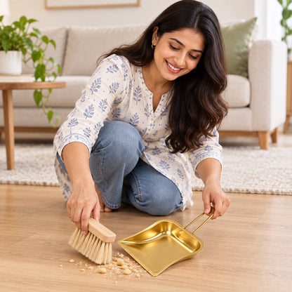 A smiling woman in a patterned shirt and jeans sweeps crumbs into a gold-tone dustpan with a wooden brush on a light floor.