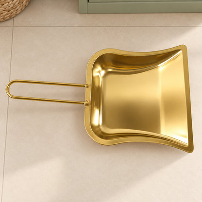 A golden metal dustpan with a loop handle lies flat on a light tiled floor, casting a soft shadow.