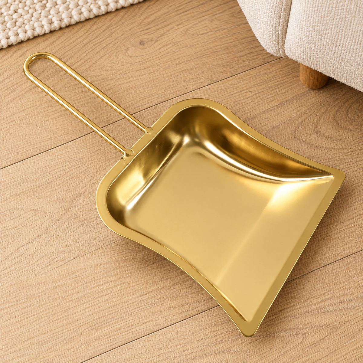 A shiny gold-tone metal dustpan with a curved loop handle rests on a light wooden floor next to a cream rug.