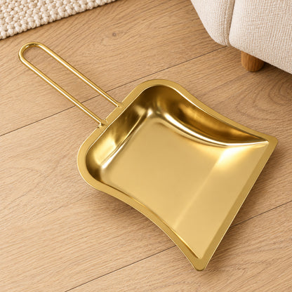A shiny gold-tone metal dustpan with a curved loop handle rests on a light wooden floor next to a cream rug.