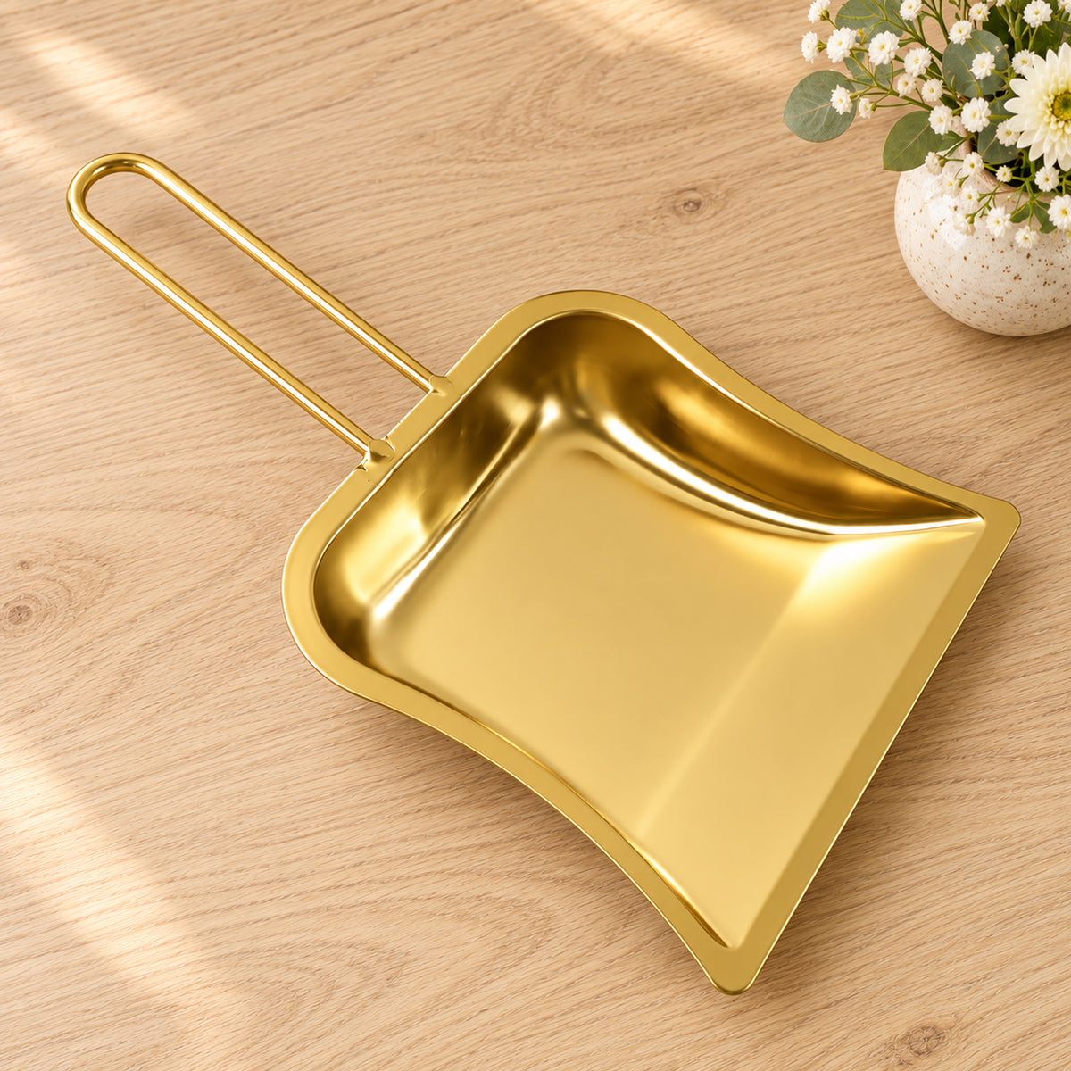 A shiny gold-toned metal dustpan with a loop handle rests on a light wooden surface next to a small white vase of flowers.