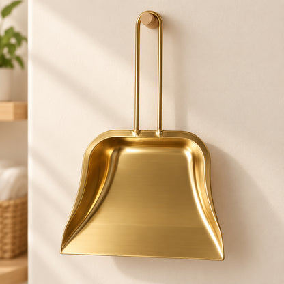 A gold-tone metal dustpan with a tall loop handle hangs on a light wooden peg against a cream wall, sunlight streaming.