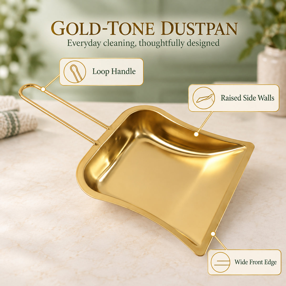 A gold-tone dustpan with a loop handle, raised side walls, and a wide front edge is displayed on a light countertop.