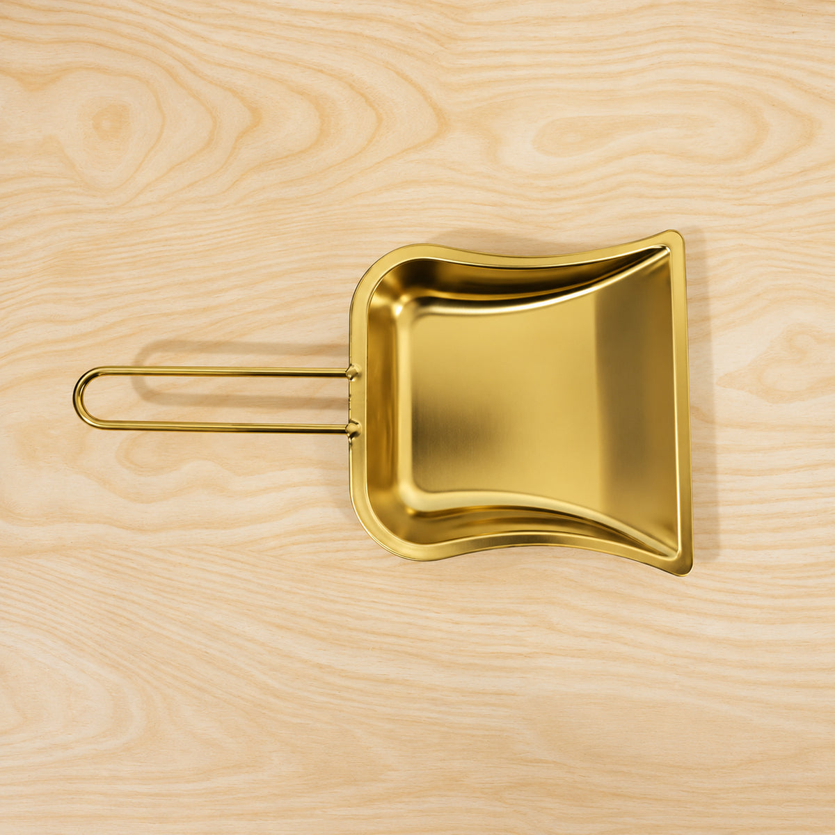 A gold-tone metal dustpan with a loop handle is positioned flat on a light-colored wooden surface with visible grain.