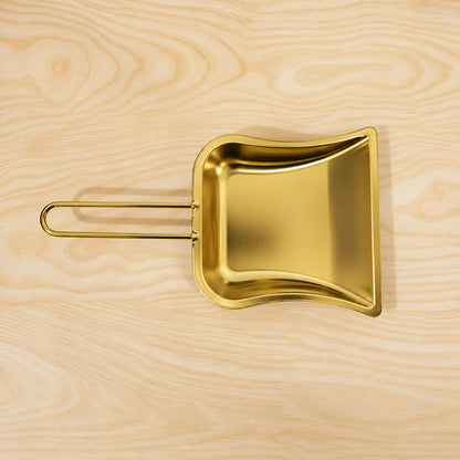 A golden metal dustpan with a loop handle is positioned horizontally on a light, finely-grained wooden surface.