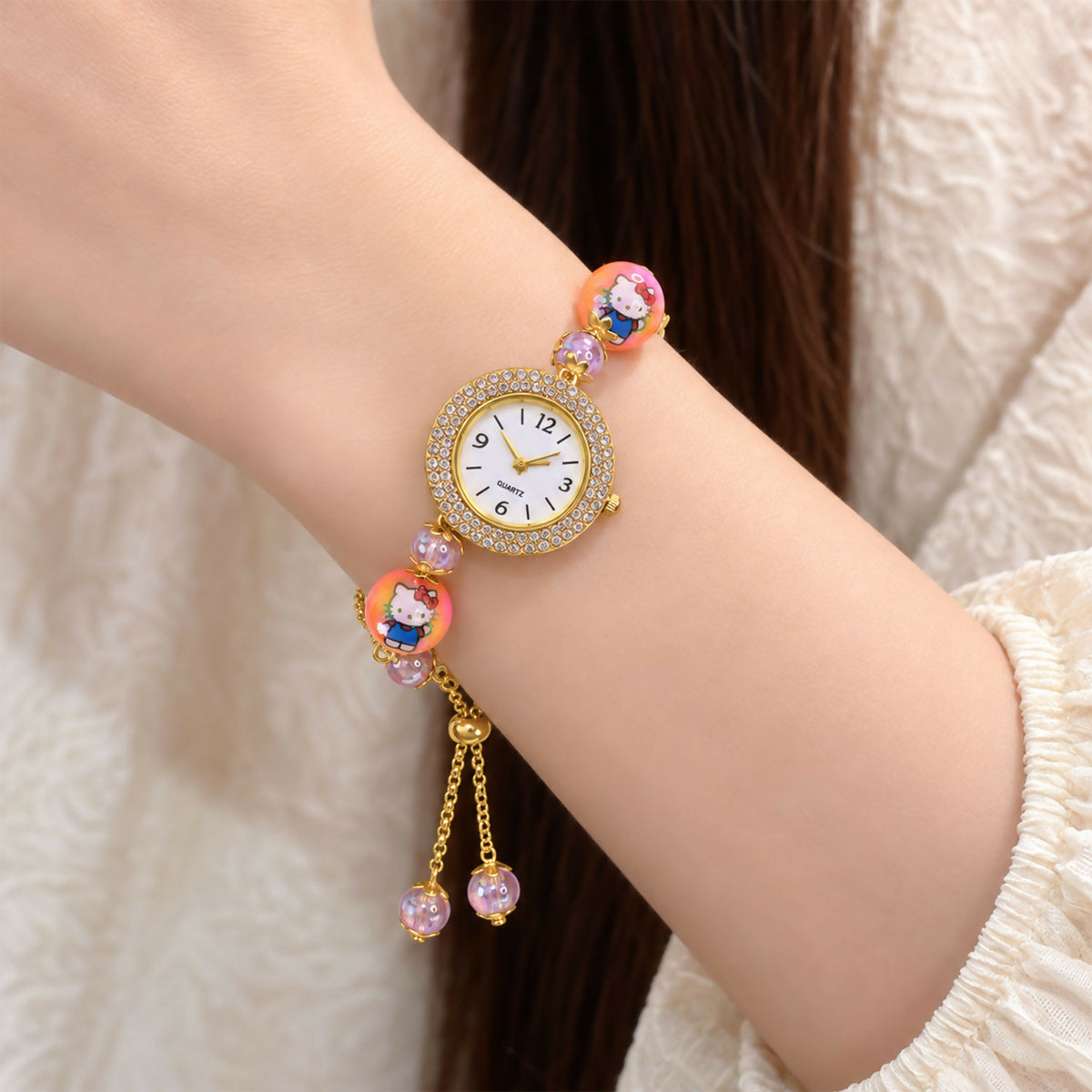 A woman's wrist wearing a gold-tone bracelet watch with a rhinestone dial, two orange cartoon character beads, and smaller