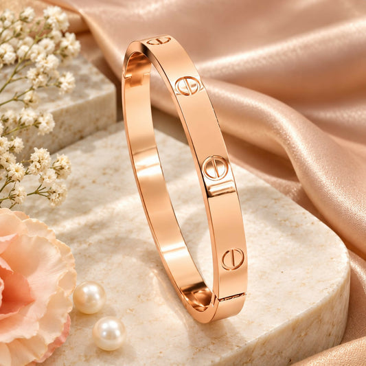 Rose gold-plated kada bracelet with engraved screw motifs resting on a light-colored stone surface with flowers and pearls.