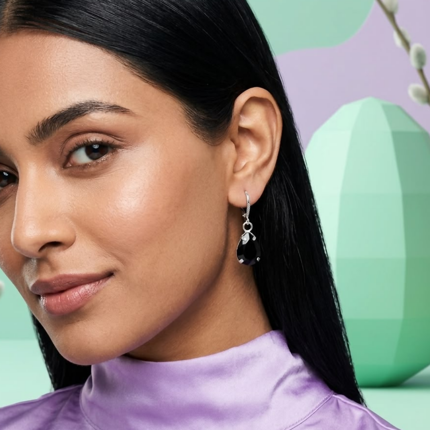 A woman with dark hair wearing a purple top and a single black teardrop crystal earring, looking towards the viewer.