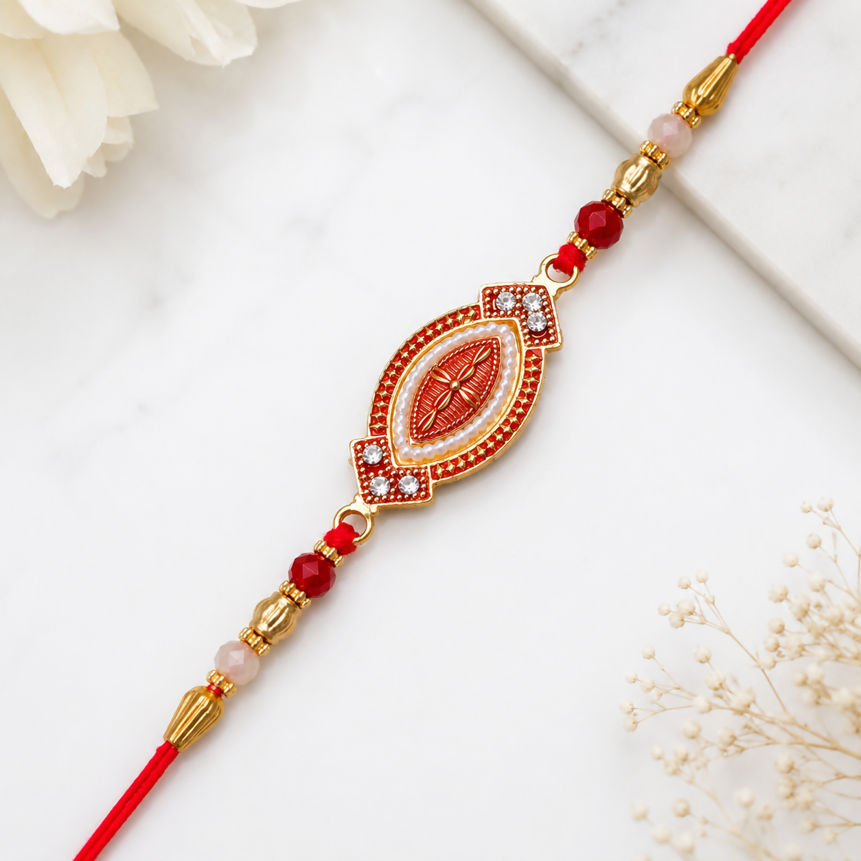 A red and gold bracelet with an oval centerpiece, adorned with white beads and red stones, lies on a white marble surface