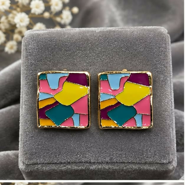 Two square stud earrings, featuring a vibrant abstract pattern, are displayed upright on a grey velvet jewelry box.