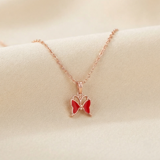 Rose gold chain with a red enamel butterfly pendant, featuring intricate wing details, resting on a beige fabric.