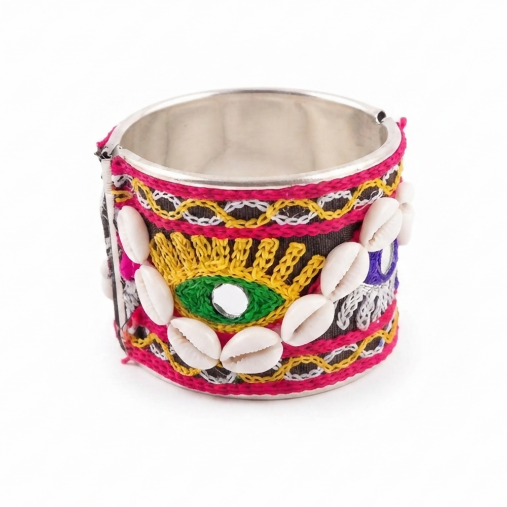 A wide silver cuff bangle with black fabric, vibrant embroidery, and cowrie shells on a white background.