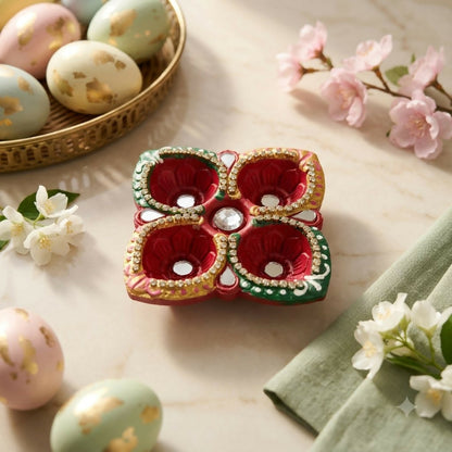A red, green, and gold four-cup diya with mirror and stone accents sits on a marble surface with decorative eggs and pink