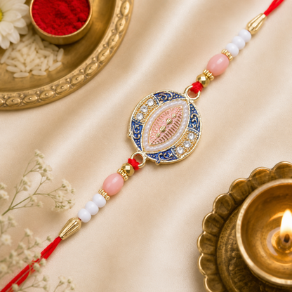 A blue, pink, and gold Rakhi with a central oval design, surrounded by small white stones and pearls, rests on a gold satin