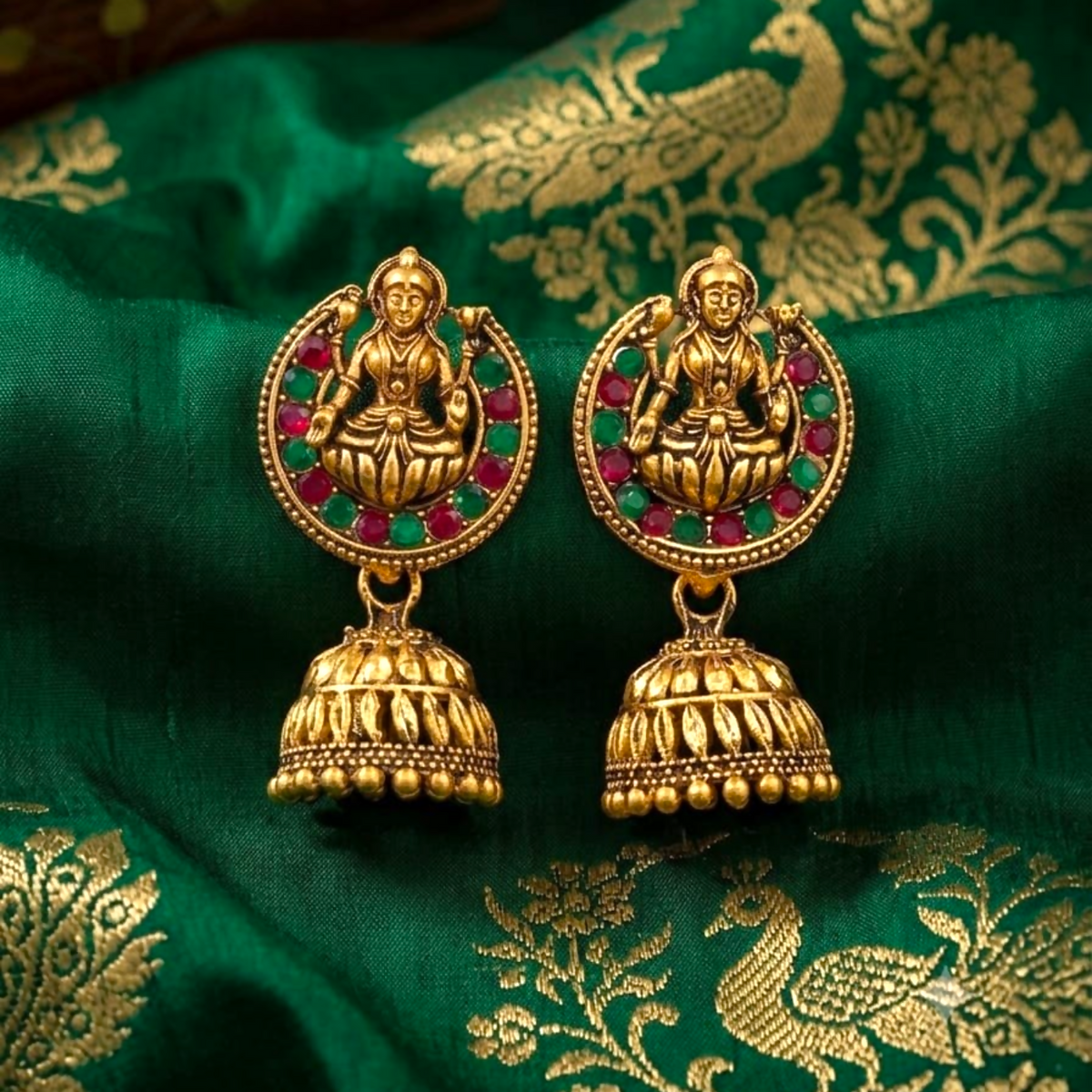 A pair of gold-toned jhumka earrings with a goddess figure and red and green stones on a green fabric.